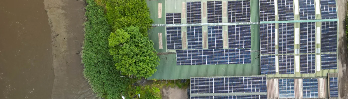 Aerial view of a riverside industrial site with solar-panel-topped buildings, various vehicles, and equipment scattered around. In the background, a bridge spans the river and mountains rise majestically, while some sustainable garden buildings add a touch of greenery to the scene.